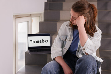 Cyber bullying. Upset teenage girl and laptop with text You Loser on stairs indoors