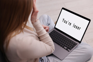 Cyber bullying. Upset teenage girl looking at laptop with text You Loser on floor indoors, closeup