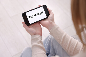 Cyber bullying. Upset teenage girl looking at smartphone with text You Loser at home, closeup