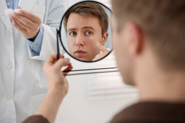 Young man putting on contact lenses near mirror in ophthalmologist office, selective focus
