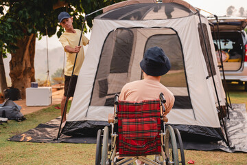 A young man with disability in wheelchair look at asian adult man setting up a camping tent, He...