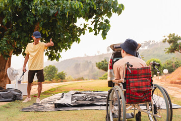 A young man with disability in wheelchair look at asian adult man setting up a camping tent, He...