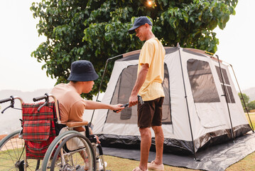 A young man with disability in wheelchair look at asian adult man setting up a camping tent, He...