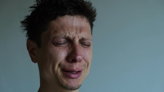 Young man talking and crying with eyes closed. Emotional close-up. Sadness, pain, inner struggle on neutral background
