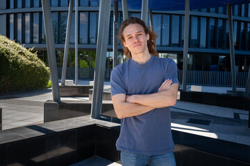 Confident young man with curly hair standing with arms crossed in modern urban environment. Outdoor...