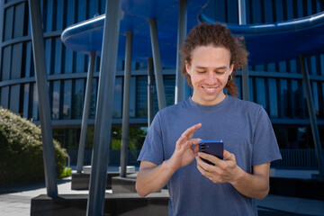 Happy smilie Young man with curly hair using smartphone outdoors in modern urban setting. Concept of digital lifestyle, technology, and communication.