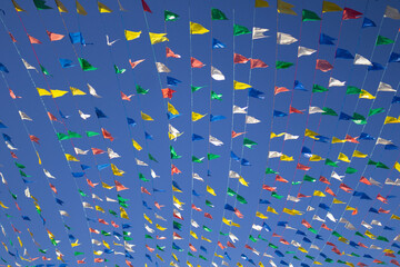 Foto de varais com bandeirolas coloridas penduradas balançando ao vento com o céu azul ao fundo. Decoração para festa junina.