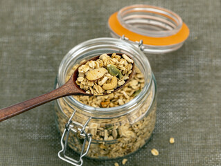 Glass jar filled with homemade granola sits on textured table, with wooden spoon lifting portion of...