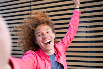 Selfie of joyful young man with curly hair and pink shirt celebrating with arms raised in front of...