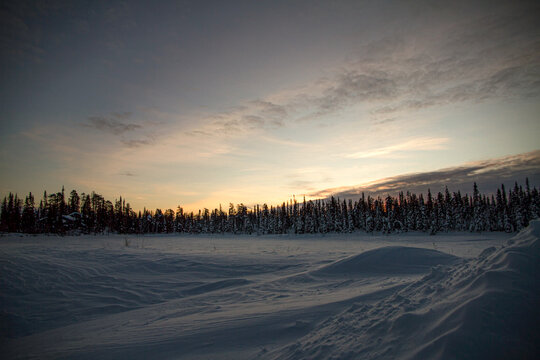 A winter road in Lapland, starting in the foreground and disappearing into the snowy forest on the horizon, where the sun sets in the spruce trees.