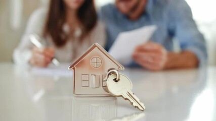House shaped keychain with key on the table close up. Couple of happy young clients signing mortgage agreement in bank office. Concept of home loan, real estate and buying own property - Powered by Adobe