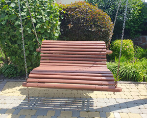 A wooden garden swing on metal chains in a sunny yard, surrounded by green bushes and ornamental...
