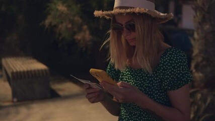 Blonde traveler checking smartphone app while counting cash, wearing sunglasses and straw hat during sunny vacation trip. Travel, blogging, weekend, relax and lifestyle concept