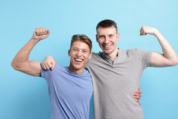 Two friends hugging and showing their biceps on light blue background