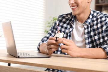 Birthday celebration. Man with cupcake having video call via laptop at table indoors, closeup