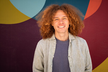 Portrait of smiling young man with curly hair standing in front of vibrant colorful mural. Urban style, relaxed expression, casual outfit, youth culture, creativity and artistic background.