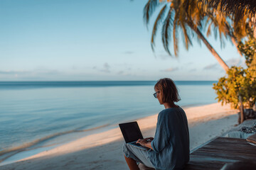 man working remotely on laptop by tropical beach during sunset with palm trees and calm sea in warm natural light