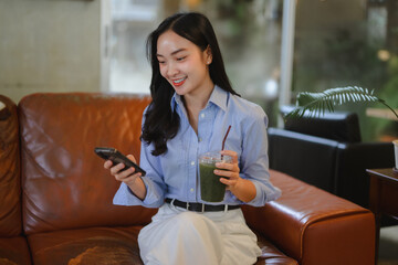Young asian businesswoman using mobile phone and drinking healthy green vegetable smoothie while...