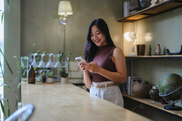 Smiling young Asia businesswoman using smartphone in modern office with wooden counter and shelves
