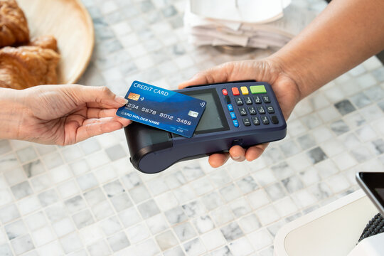 Two adult hands exchanging payment using blue credit card with wireless symbol on electronic POS terminal placed on marble table surface in cafe environment with croissant plate in background