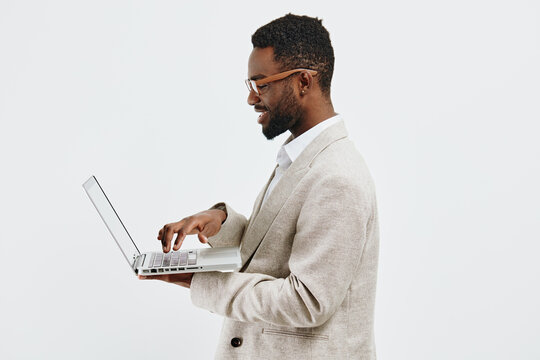 Professional young African-American man in stylish beige blazer using silver laptop computer, holding and typing on it while smiling, isolated on plain white background, studio portrait, business - Powered by Adobe