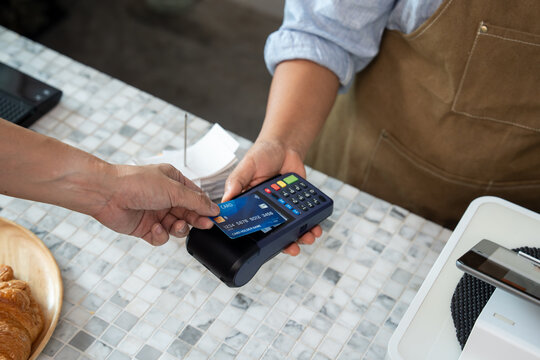 Customer hand tapping blue credit card with chip and wireless sign onto POS terminal held by barista wearing brown apron over marble table counter in cafe during contactless payment process