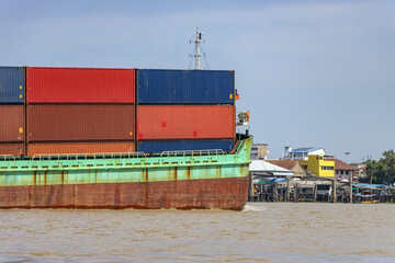 Cargo ship carrying stacked containers on a river near city waterfront, Thailand