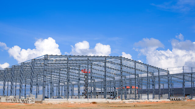 Metal structure of large factory warehouse industry buildings with construction workers are working in construction site against clouds on blue sky background