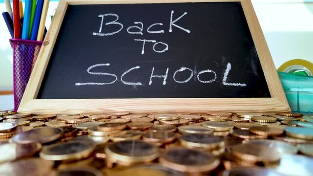 Euro coins scattered on a table with a small blackboard with the words back to school written on it and colored pencils and adhesive tape in the background