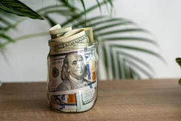 Glass jar full of rolled and stacked US hundred dollar bills on wooden table with green tropical...