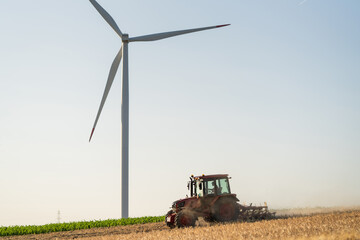 Agricultural tractor is working on the field with wind turbines