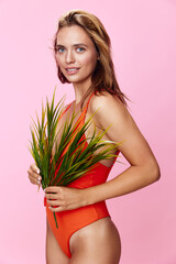 Vibrant woman in orange one piece swimsuit posing with plant against a soft pink background