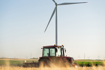 Agricultural tractor is working on the field with wind turbines