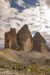 Iconic mountain peaks known as the Tre Cime di Lavaredo rise above a rocky valley in the Italian Alps. Dramatic clouds and rugged terrain evoke a sense of wilderness and grandeur.