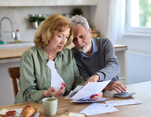 Senior couple checking bills and calculating expenses using a calculator at home, debt taxes and home budget concept, worried married elderly couple