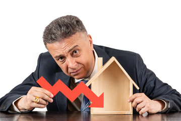 Middle aged man in business suit holding red arrow falling graph in hands and miniature wooden...