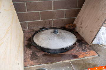 Large cauldron with a steaming lid simmers in a brick oven built into the wall with wood paneling on the sides, creating a cozy and rustic cooking space.