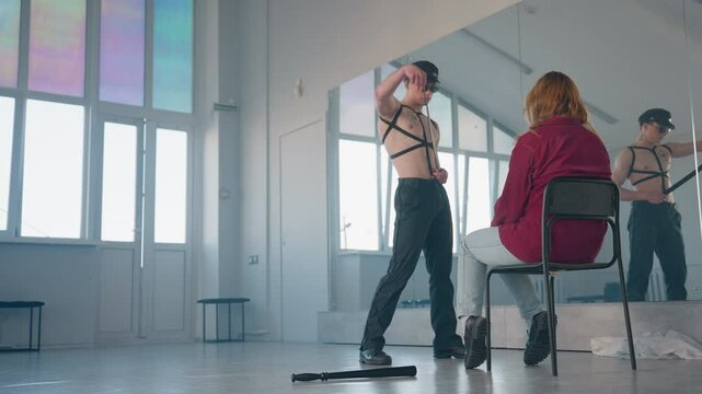 man removes belt then wraps strap around neck and tosses it aside in front of woman seated on chair in bright mirrored dance studio with baton and soft cloth on floor near reflection