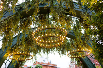 A decorative arch decorated with lush greenery and round illuminated chandeliers.