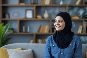 Close-up portrait of a young Muslim woman in a hijab and shirt sitting on a sofa at home, smiling...