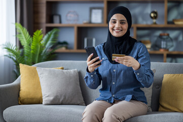 Smiling young Muslim woman in hijab sitting on sofa at home, holding credit card and phone, looking at camera