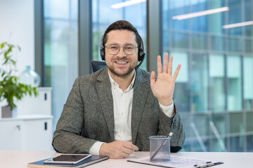 Portrait of a businessman in a headset and suit sitting at a desk in the office, smiling and waving at the camera