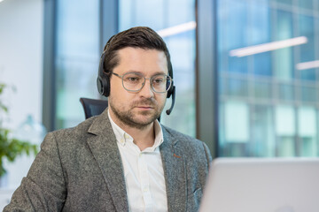 Close-up photo of a young man in a suit and headset working seriously in the office on a laptop