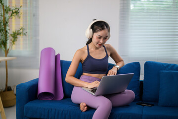 Obraz premium Asian woman wearing headphones using laptop sitting on sofa with yoga mat after workout at home