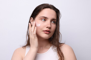 Beautiful woman applying skincare product onto her face on light grey background, closeup