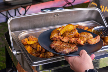 Person is holding a plate of food, which includes meat and potatoes. The plate is placed on a silver tray, and the person is reaching for it. The scene suggests a casual and relaxed atmosphere.