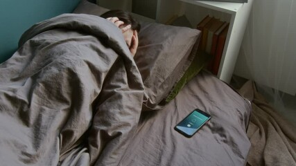 Shot of Caucasian teen girl waking up in morning by loud smartphone alarm switching it off and hiding under bed covers - Powered by Adobe