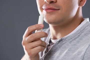 Young man applying lip balm onto his lips on grey background, closeup