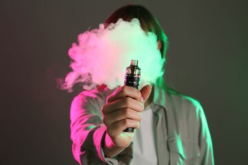 Woman using electronic smoking device in neon lights on grey background, selective focus