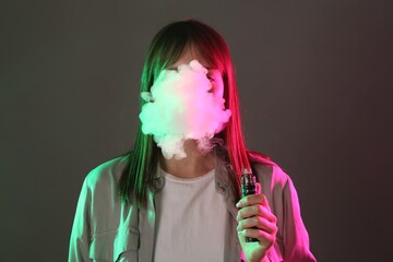 Woman using electronic smoking device in neon lights on grey background